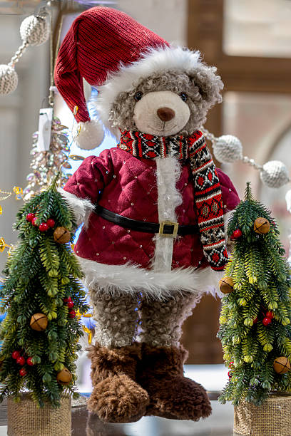 istock Christmas decoration plush bear in red Santa-style jacket, knitted hat, and patterned scarf, standing between two small Christmas trees 2244782947