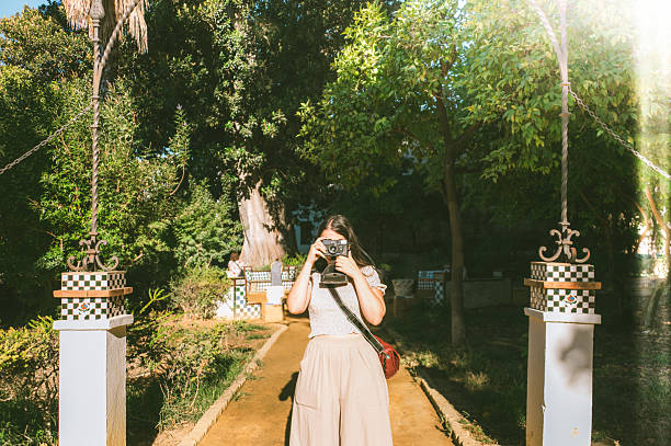 A young tourist woman in a public park of Seville, Spain, taking a photo using a vintage photographic film camera.