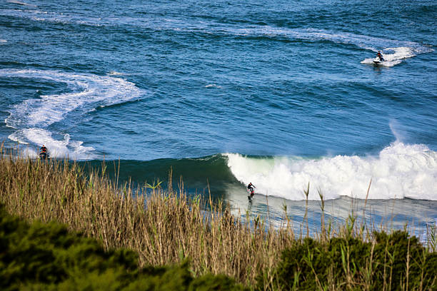 A surfer rides a breaking wave while two people on jet skis wait in the ocean further offshore. The scene is viewed from land with coastal vegetation in the foreground. Blue open water and white spray fill the frame on a sunny day.
