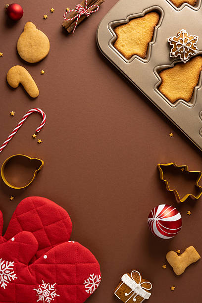 istock Festive top view vertical Christmas baking scene featuring a pan of tree shaped cookies, candy cane, mitten, ornaments and gift accents on a warm brown background 2244794252