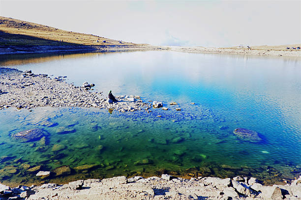 A peaceful moment by a high-altitude lake, where calm water reflects the sky and mountains. The figure sitting on the rocky shore adds a sense of solitude and contemplation. Captured in natural light in the highlands of Turkey.