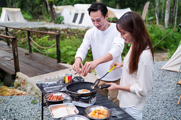 The lovely Asian couple embarks on an outdoor cooking adventure and preparing delicious meals together near to their campsite during weekend holiday vacation at a natural parkland.