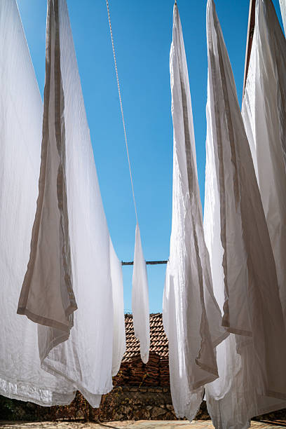 Washing day under bright blue sky. Crisp white sheets billow gently in the sun, creating dramatic lines against rustic terracotta roof and stone wall below.