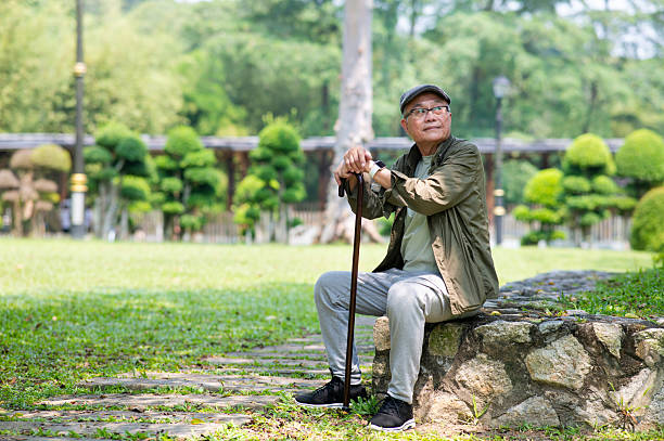 A comfortable spring daytime park with plenty of greenery in Japan. A Japanese man in his late 70s is sitting on a bench wearing a suit. He holds a book and looks into the distance.