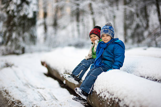Two little boys dressed in warm winter clothing sit on large snow-covered logs in a forest during winter. The children smile at the camera, while snow blankets the ground and trees. The scene captures a quiet moment of outdoor play or rest in the natural beauty of a snowy landscape.Shot with Nikon D800