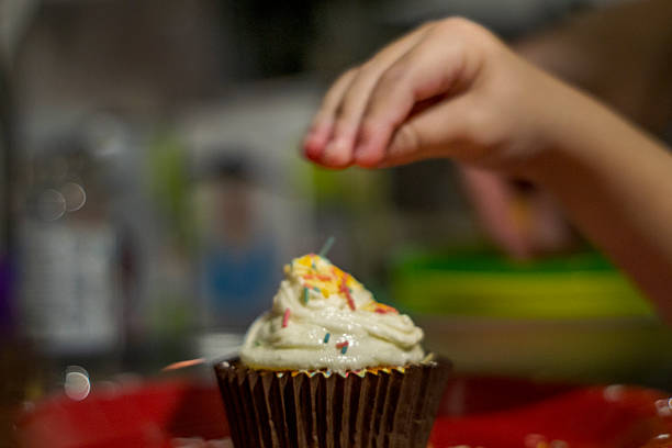 A chocolate cupcake topped with white frosting and colorful sprinkles sits on a red surface. A child’s hand hovers above the cupcake, either reaching for the treat or adding sprinkles, capturing a candid moment of interaction. The blurred background and shallow depth of field draw attention to the gesture and dessert, evoking themes of celebration, curiosity, and joyful engagement.