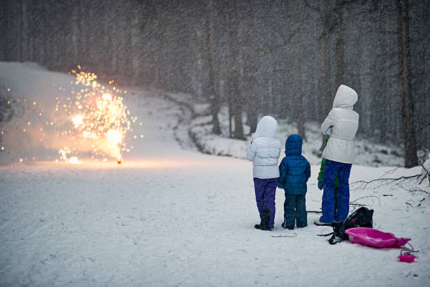 Three children dressed in warm winter clothing stand together in a snowy outdoor setting, watching colorful fireworks light up the winter road. The scene captures a joyful winter celebration in a forested area, highlighting the contrast between the cold, quiet landscape and the vibrant bursts of fireworks. Shot with Nikon D800