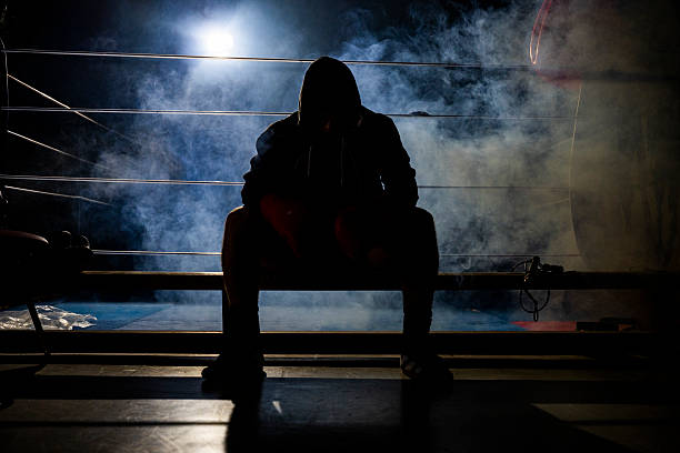 Focused young male boxer preparing mentally inside a dimly lit boxing ring, surrounded by a smoky atmosphere. The setting conveys a sense of intensity and concentration as he gets ready for his next challenge.