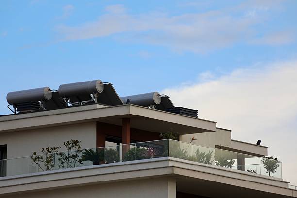 Solar thermal water heating systems mounted on the rooftop of a modern apartment building, with a large balcony planted with ornamental greenery and a partly cloudy blue sky above. Renewable energy technology on contemporary residential architecture.