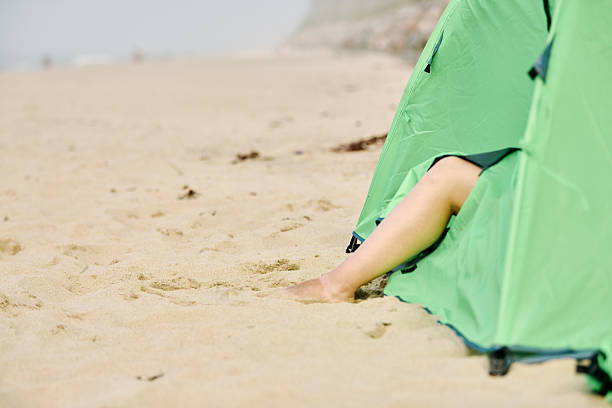 Serene beach setting features green tent pitched on golden sand with persons feet peeking out. Soft lighting and distant sea waves create calming atmosphere ideal for coastal tourism and leisure