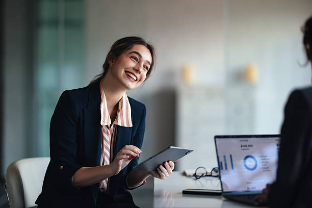 istock Smiling Professional Woman in Office Discussing Tablet While Laptop Displays Data Charts 2244825262