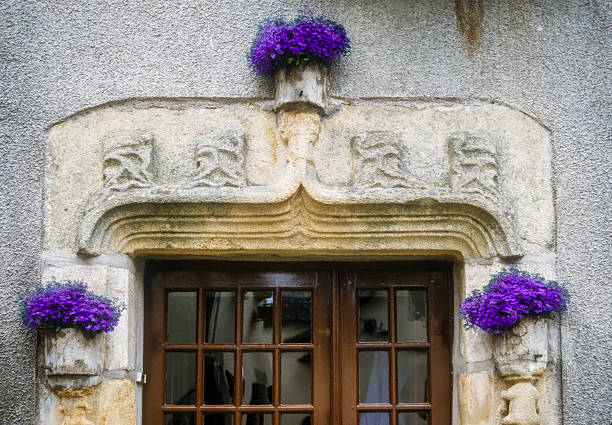 Dark wooden window framed by a carved stone arch. Three stone flowerpots are placed above and on either side of the window, each containing bright purple flowers