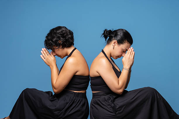 Two women dressed in black sitting back-to-back on chairs against a blue background, leaning forward with their palms together in a prayer gesture, symbolizing hope, meditation, or gratitude.