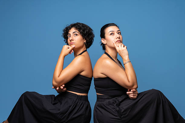 Studio portrait of two women sitting back to back with thoughtful and distant gazes, looking away from each other.