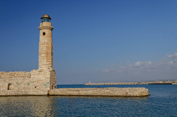 A serene coastal scene featuring the historic stone lighthouse of Rethymno, Crete, Greece. Positioned on a pier that stretches into calm blue waters, the cylindrical tower stands under a clear sky, its lantern room crowning the structure. A long breakwater extends into the distance, adding depth to the tranquil maritime setting. This image captures the elegance and functionality of traditional coastal architecture in a picturesque Mediterranean port.