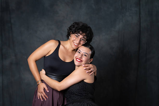 Two cheerful young women smiling and embracing each other while posing in front of a dark backdrop.