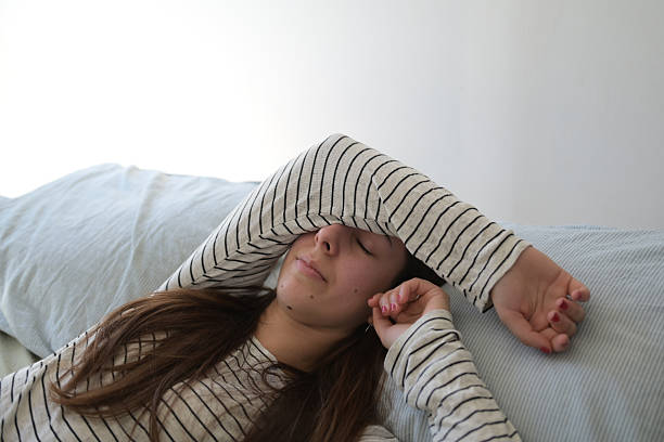 Young woman with sleep mask waking up in bed and snoozing alarm clock on cell phone, mobile telephone. Girl sleeping in bedroom at home and waking up after receiving text message or call on smartphone