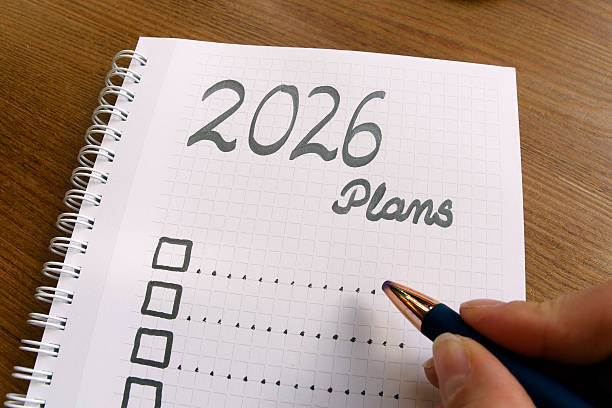 istock 2026 Plans,  A spiral notebook on wooden desk with checkboxes for goals, planning, and future ideas for upcoming New Year., with grid pages and hand holding blue pen 2244847177