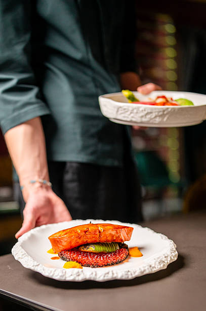 A professional chef plates a delicious seared salmon dish with a green vegetable and orange garnish in a high-end restaurant setting. The chef's hands are out of focus in the background
