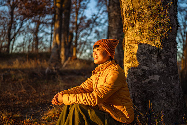 Woman wearing a knit hat and a yellow jacket sitting against a tree, relaxing and smiling in a forest during a golden hour sunset, experiencing peace and connection with nature