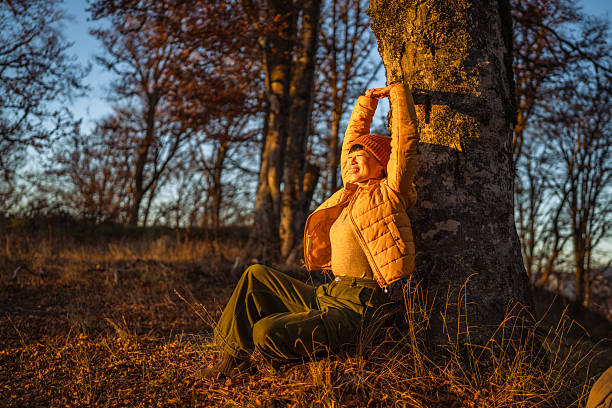 Young woman leaning against a tree trunk in an autumn forest, stretching her arms above her head and smiling with closed eyes, feeling refreshed during golden hour