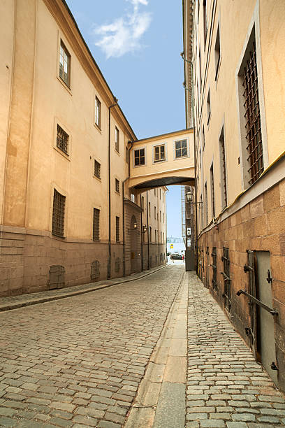 A cobbled alley flanked by historic beige-brown stone buildings, featuring an overhead enclosed walkway. The view is bordered by intricate architectural details, under a clear sunny sky. Stockholm, Sweden