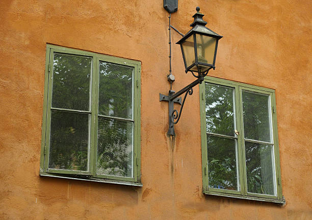 A beautifully textured ochre wall contrasting against a pair of green wooden-framed glass windows. The windows reflect lush greenery outside, complemented by a black wrought iron lantern affixed to the wall. The old city of Stockholm, Sweden