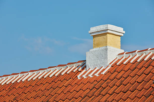 A detailed view of a traditional roof with red clay tiles and a white masonry chimney, set against a serene blue sky. The scene evokes simplicity and timeless architectural techniques.