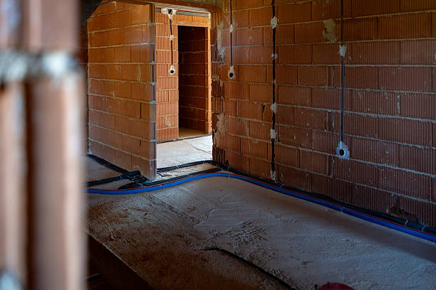 An indoor view of a house under construction shows raw red brick walls, exposed electrical conduits, and unfinished concrete flooring. Doorways lead into adjacent rooms, highlighting the early phase of the building process. The scene reflects construction work and infrastructure development.