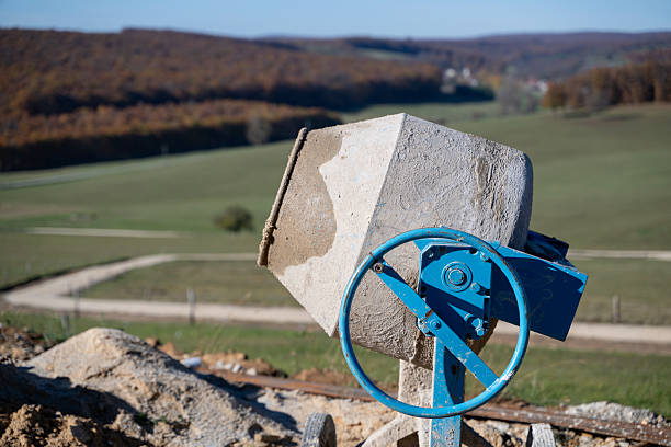 A weathered concrete mixer with a blue handwheel stands on a construction site overlooking rolling hills and open fields. The natural autumn landscape contrasts with the industrial equipment, highlighting rural development and infrastructure work in progress.