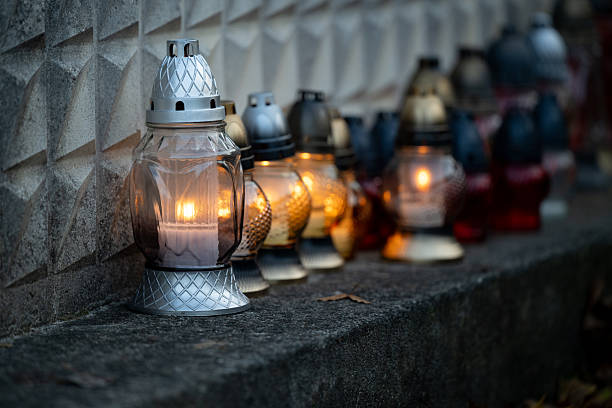 Multiple grave candles of various colors and designs are lined up along a concrete ledge in front of a textured stone wall. The flames glow warmly in the dim light, creating a solemn and respectful atmosphere. The image reflects remembrance and cultural tradition during a time of mourning.