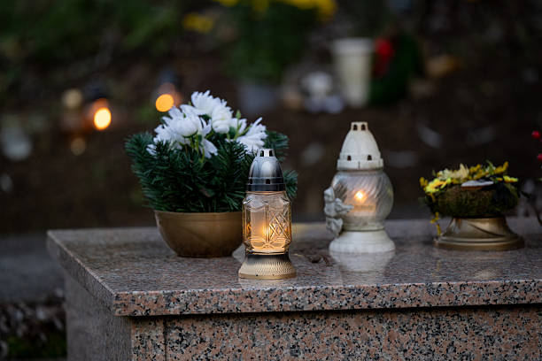 Multiple grave candles of various colors and designs are lined up along a concrete ledge in front of a textured stone wall. The flames glow warmly in the dim light, creating a solemn and respectful atmosphere. The image reflects remembrance and cultural tradition during a time of mourning.