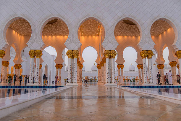 Colonnade with golden arches and reflection pool at Sheikh Zayed Grand Mosque, a major cultural and touristic site in Abu Dhabi, at sunset. Publicly available place, no tickets required.