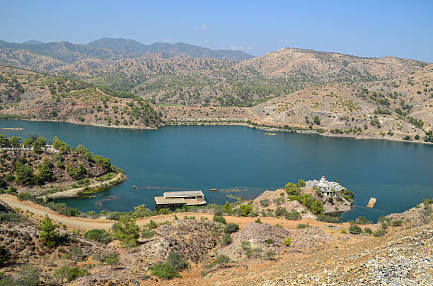A panoramic view of the Xeros River reservoir (likely Asprokremmos Dam) surrounded by arid hills, featuring a submerged or abandoned stone structure on a small island, illustrating a peaceful, yet complex water management landscape.