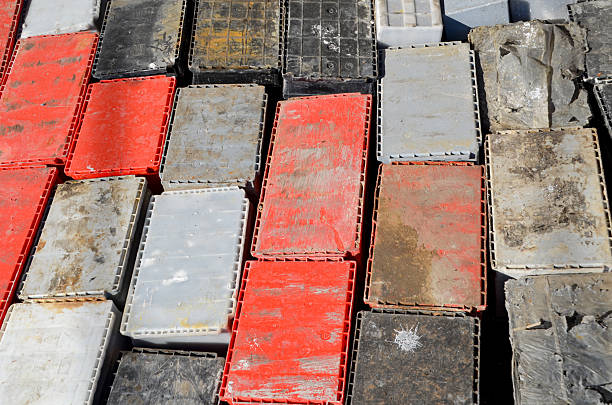 A high-angle view of rows of used lead-acid car battery casings, featuring red, gray, and black colors, stacked and collected for recycling. The dirty, industrial scene highlights electronic and hazardous waste.