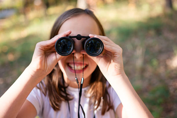 A sweet little brunette haired girl stands among some large sawed off tree trunks, as she holds a pair of binoculars to her eyes and looks out.  She is dressed casually in layers to stay warm and has a baseball hat on backwards.