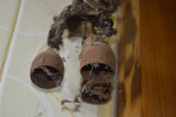 rusty metal bells hang from a frayed rope, their surfaces eroded and layered with dust and spiderwebs. A neglected, vintage look evokes time, decay, and quiet, forgotten corners of a home.