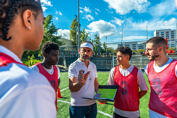 Soccer coach with clipboard explains tactics to a focused group of young adult male players during sunny practice on artificial turf, emphasizing teamwork and strategy