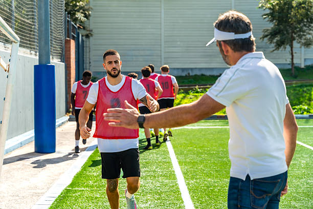 Soccer team members running on a training pitch, with a coach extending his hand towards a player, symbolizing guidance, support, and teamwork during practice