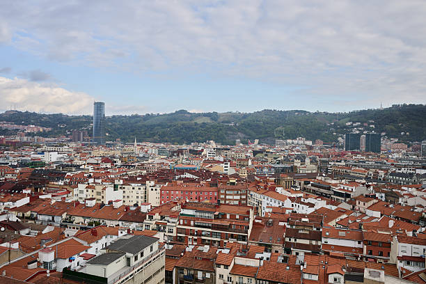 Bilbao cityscape featuring red tile rooftops, modern buildings, and the surrounding green hills under a cloudy sky