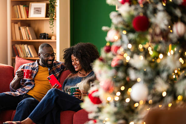 istock Cheerful black couple using phone while relaxing on sofa at home on Christmas holidays 2244920492