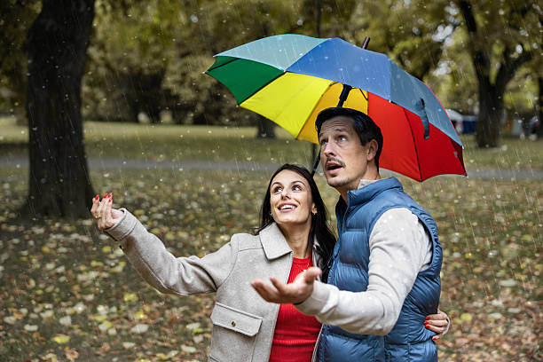 A joyful couple shares a vibrant umbrella while enjoying a rainy day, expressing happiness and spontaneity amidst falling raindrops in a picturesque park environment.