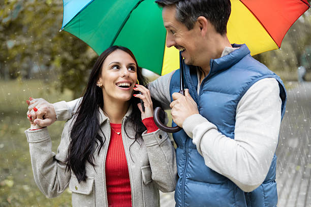 Young adult Black woman and young adult Caucasian man walking together under transparent umbrella on rainy street, smiling and talking, holding hands, illuminated brick building in background
