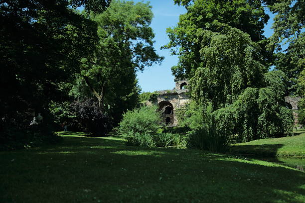 Lush green park landscape with old stone ruins. Peaceful park scene with dense green trees surrounding ancient stone ruins under a bright summer sky. The sunlight creates soft shadows across the grass highlighting the calm atmosphere of the natural landscape. The mix of nature and historical architecture forms a beautiful composition showing harmony between human history and the living environment in a tranquil garden setting.