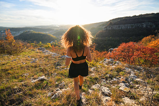A mid-adult woman with curly hair runs along a trail in a peaceful fall landscape, focusing on her workout and wellness.