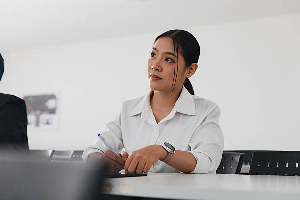 A focused young woman sits at a desk in a bright classroom, writing notes. She looks attentive, wearing a white shirt and holding a pen. Other students can be seen nearby.