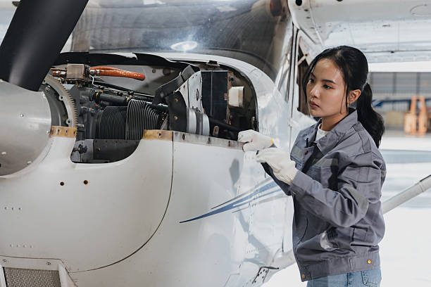 A young woman in a gray jacket carefully inspects the engine of a small aircraft inside a hangar. She is focused on the task while wearing gloves for safety.