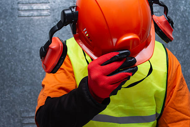 istock Orange protective safety helmet with ear muffs held by a worker wearing a reflective vest. Concept of industrial safety equipment and protection at work. 2244944948