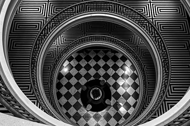 An overhead view of a grand spiral staircase showcasing perfect symmetry and geometric precision. The contrasting black-and-white tones emphasize the bold lines, circular motion, and checkered marble floor, creating a visually captivating architectural composition.