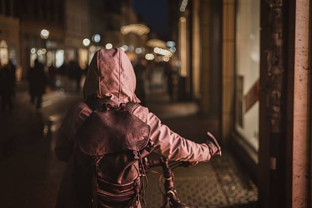 A rear view of a person wearing a hood and backpack cycling down a cobblestone street at night, conveying urban solitude and travel.